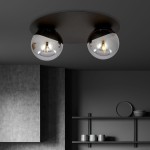 Czarna/grafitowa lampa sufitowa 2-punktowa z dwiema szklanymi, okrągłymi kloszami. Ujęcie z dołu, lampy zamontowane na okrągłej podstawie. Widoczne metalowe detale oraz odbicia światła w kloszach, w tle szare ściany i półki z naczyniami.