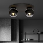 Solar lampa sufitowa 2-punktowa w kolorze czarnym z okrągłą podstawą. Dwa szklane klosze z transparentnym wykończeniem, z widocznymi żarówkami. Lampa zamontowana na szarym tle, w minimalistycznym wnętrzu z półkami i ceramicznymi naczyniami.