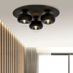 Czarna lampa sufitowa 3-punktowa z okrągłymi kloszami, z przezroczystymi elementami, zawieszona na okrągłej podstawie. Klosze mają widoczne żarówki, a tło to gładki sufit. Lampa pasuje do nowoczesnych wnętrz.