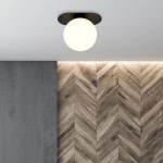 Lampa sufitowa Plaza 1-punktowa w kolorze czarnym i białym, z okrągłym, mlecznobiałym kloszem. Zainstalowana na białym suficie, w otoczeniu ściany z teksturą betonu i drewnianych paneli w układzie jodełkowym.