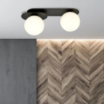 Czarna lampa sufitowa Plaza 2-punktowa z białymi, okrągłymi kloszami. Ujęcie z dołu, montowana na suficie. Tło z szarej ściany i drewnianych paneli w układzie jodełkowym.