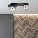Lampa sufitowa Plaza 2-punktowa w kolorze czarnym i grafitowym z dwoma szklanymi kulami. Montowana na suficie, z widocznymi elementami metalowymi. Tło w odcieniach szarości i drewna, tworzy nowoczesną aranżację.