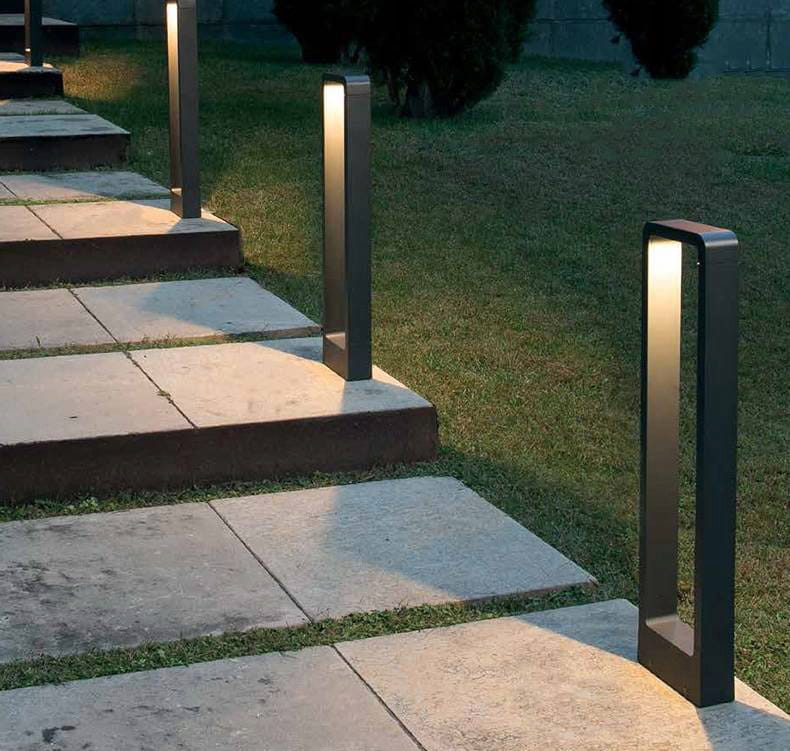 Lampa stojąca ogrodowa Rio LED H80 w kolorze czarnym, w kształcie prostokątnego słupka, umieszczona wzdłuż kamiennej ścieżki, emitująca światło z góry. Widoczna na tle trawnika i drzew.