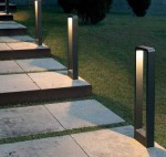 Lampa stojąca ogrodowa Rio LED H80 w kolorze czarnym, w kształcie prostokątnego słupka, umieszczona wzdłuż kamiennej ścieżki, emitująca światło z góry. Widoczna na tle trawnika i drzew.