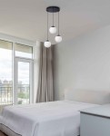 Lampa wisząca Camina 3-punktowa w kolorze czarnym, z trzema okrągłymi, białymi kloszami. Zawieszona w sypialni, z widocznym jasnym tłem ściany i oknem. Klosze umieszczone na cienkich, czarnych linkach.