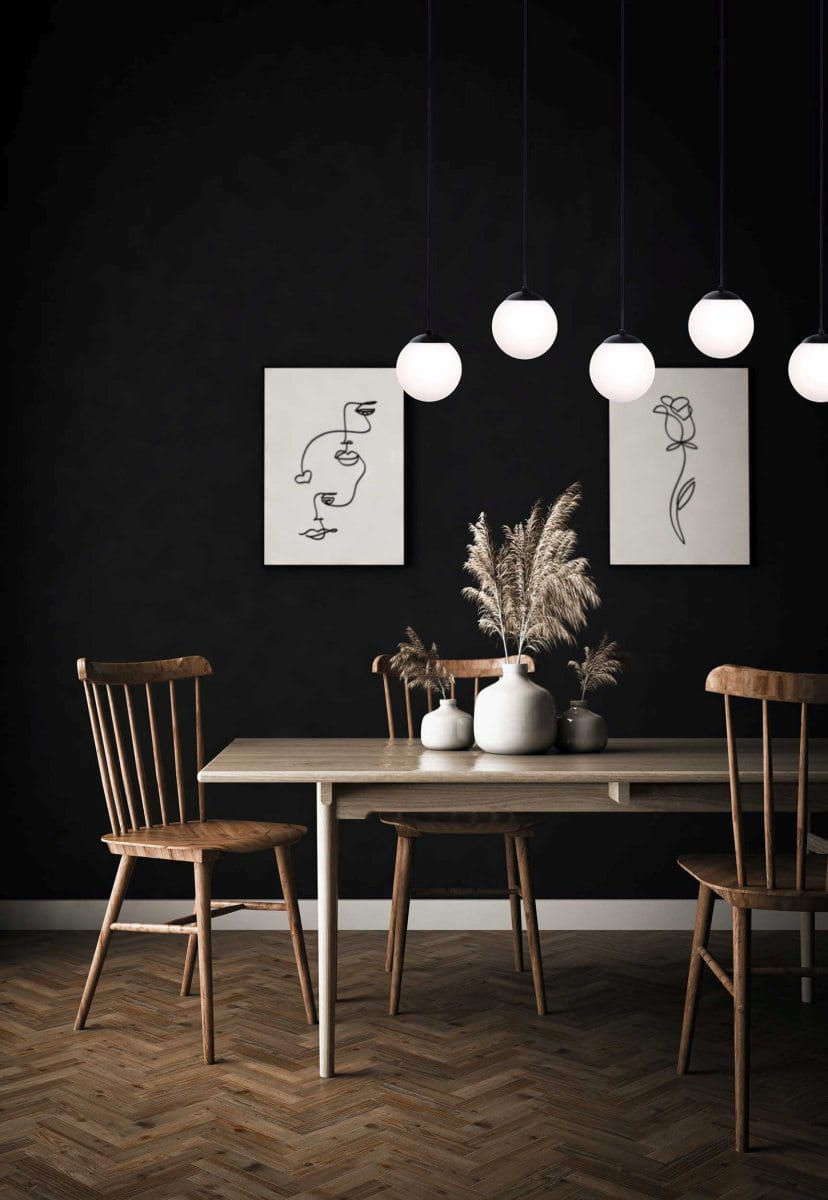Lampa wisząca Camina 5-punktowa w kolorze czarnym, z pięcioma okrągłymi kloszami w białym kolorze, zawieszona w poziomie nad drewnianym stołem. Tło w ciemnym kolorze, z dekoracjami ściennymi i roślinnymi na stole, tworzy nowoczesną aranżację wnętrza.