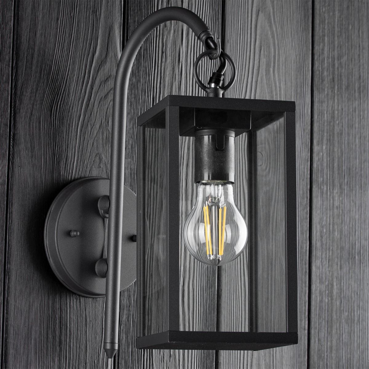 Malmo kinkiet elewacyjny ścienny 1-punktowy w kolorze grafitowym. Konstrukcja z metalowym korpusem i przezroczystym kloszem, zawieszony na zakrzywionym ramieniu. Widoczna żarówka o klasycznym kształcie, montowany na ścianie, na tle drewnianej faktury.