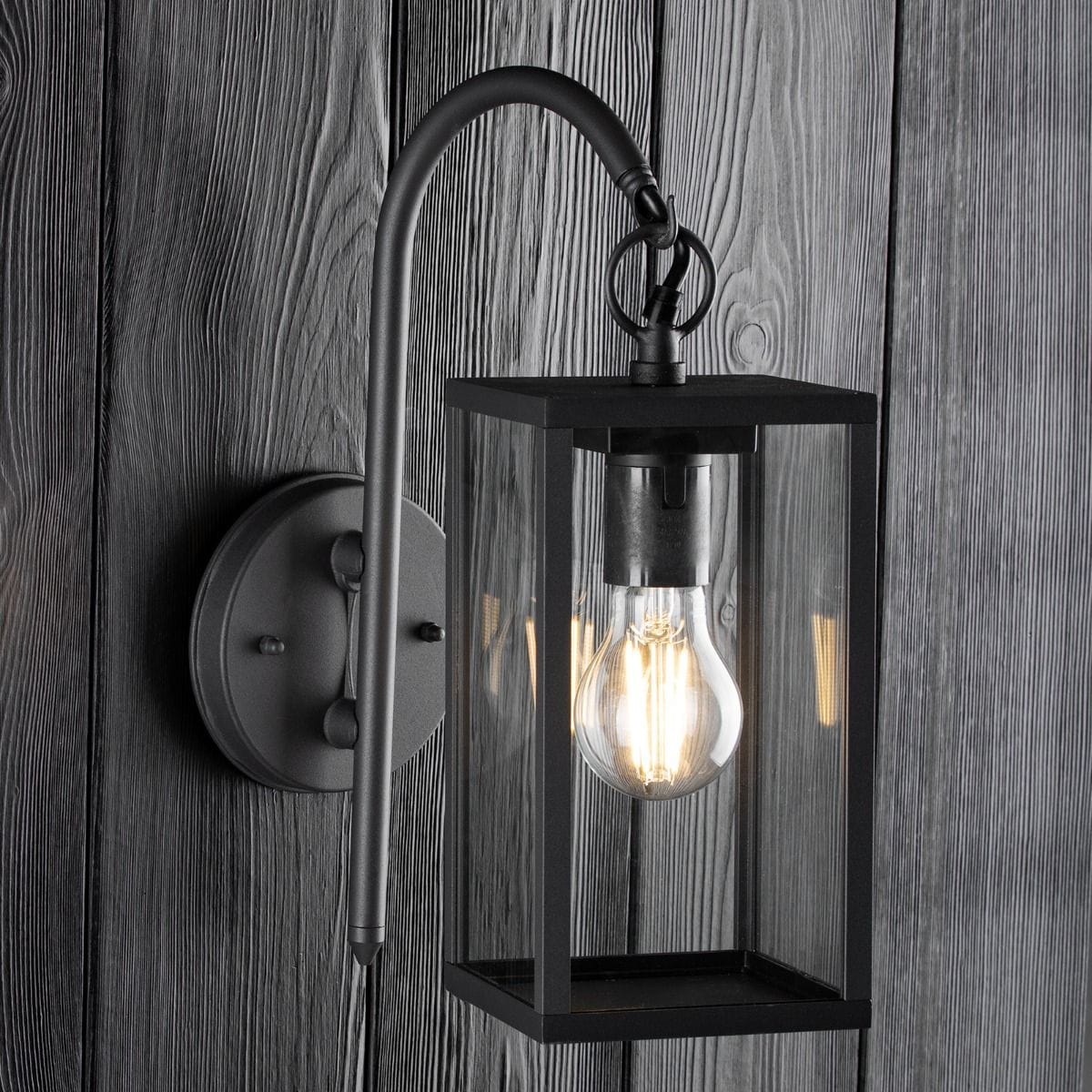 Kinkiet elewacyjny Malmo w kolorze grafitowym, 1-punktowy. Produkt ma prostokątną, szklaną obudowę z metalowym stelażem, zamontowany na ścianie, z widoczną żarówką. Ujęcie z boku, na tle drewnianej powierzchni.