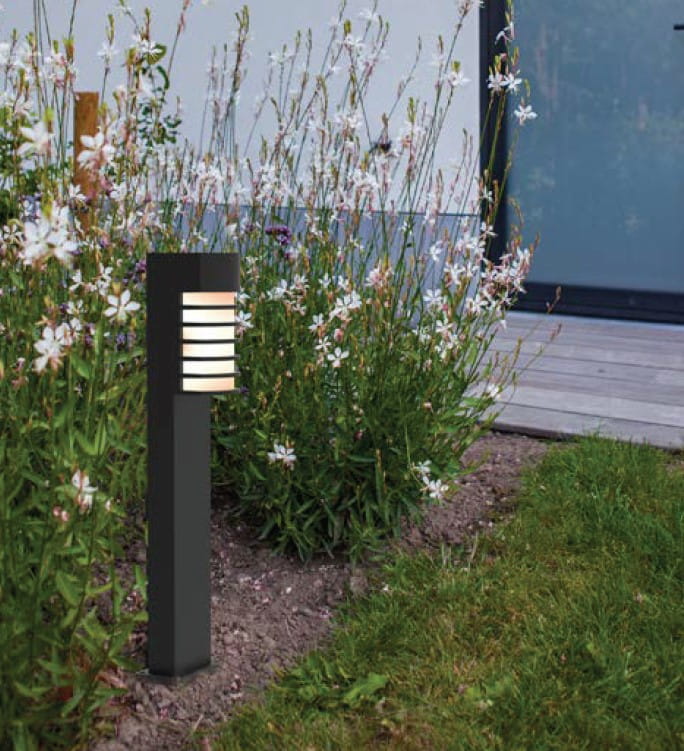 Lampa stojąca ogrodowa Fulton H75, czarna, z prostokątnym korpusem, oświetleniem w górnej części z poziomymi żebrami. Ustawiona na tle kwiatów i trawnika, w naturalnym otoczeniu.