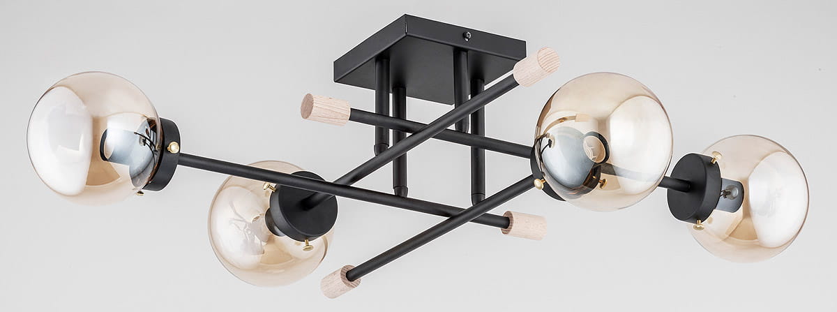 Czarna lampa sufitowa 4-punktowa z metalowymi ramionami w kształcie krzyża. Szklane klosze w odcieniu złota z widocznymi żarówkami. Elementy drewniane przy mocowaniach. Produkt zamontowany na prostokątnej podstawie.
