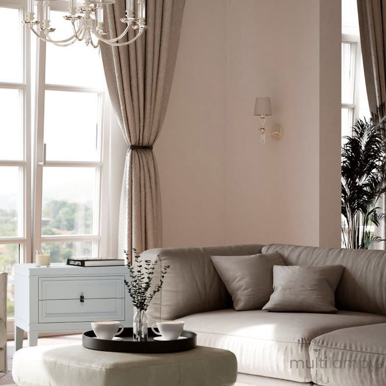 Kinkiet Siena 1-punktowy W01315WH w kolorze białym, zamontowany na ścianie w salonie, obok okna. Stylowy, minimalistyczny design z delikatnym abażurem. W tle widoczny szary narożnik, stół kawowy z filiżankami i kwiatami, oraz zasłony.