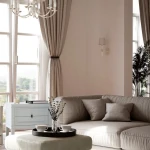 Kinkiet Siena 1-punktowy W01315WH w kolorze białym, zamontowany na ścianie w salonie, obok okna. Stylowy, minimalistyczny design z delikatnym abażurem. W tle widoczny szary narożnik, stół kawowy z filiżankami i kwiatami, oraz zasłony.