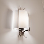 Kinkiet Faro 1-punktowy W01053NI o stalowej, połyskliwej podstawie z białym, cylindrycznym kloszem. Ujęcie z boku, montowany na ścianie, z eleganckim wykończeniem i delikatnymi detalami. Tło w jasnym kolorze.