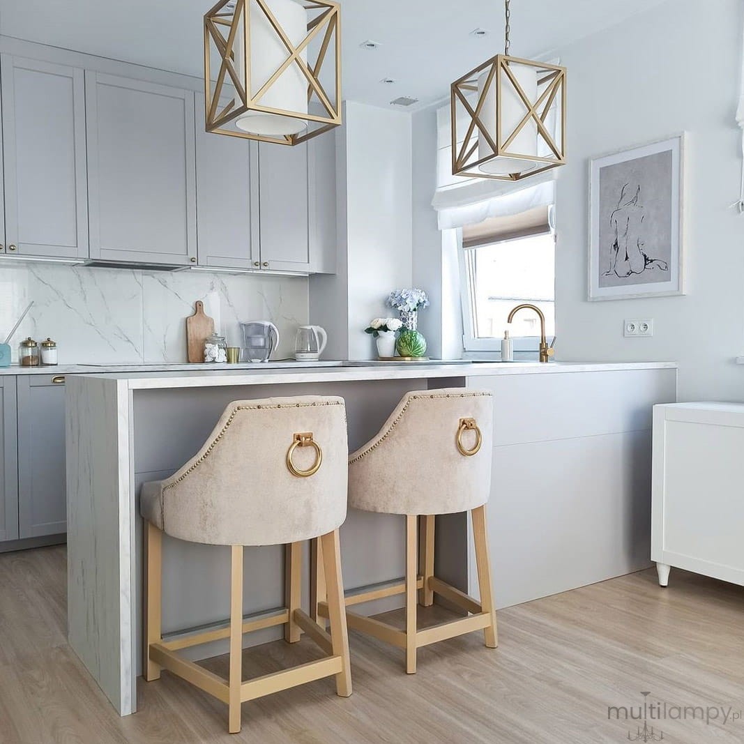 Lampa wisząca New York 1-punktowa P01403WH AU w geometrycznym kształcie, z białym kloszem i złotą ramą, zawieszona nad wyspą kuchenną. W tle nowoczesna kuchnia z szarymi szafkami i drewnianą podłogą, dwa tapicerowane stołki barowe.