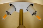 Lampa biurkowa Fungo 2-punktowa w kolorze złotym i mosiądzu. Widok z góry na dwa źródła światła w białych oprawach, z widocznymi żarówkami o pomarańczowym kolorze. Metalowa podstawa w złotym odcieniu.