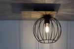 Czarna lampa sufitowa Cyber Prostokąt z dębową podstawą. Okrągła klosz w formie siatki metalowej, z widoczną żarówką. Ujęcie z dołu, montowana na suficie, w tle jasna ściana. Idealna do nowoczesnych wnętrz.