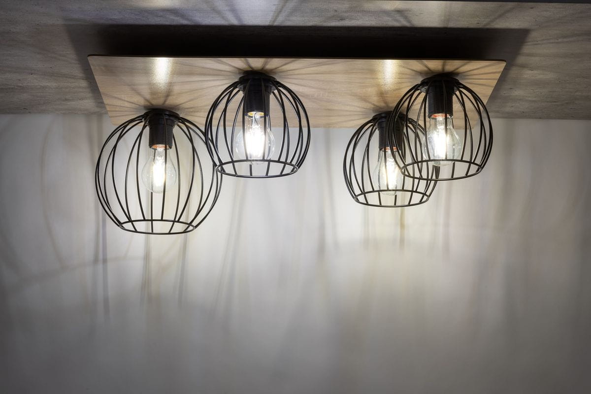 Czarna lampa sufitowa 4-punktowa z oprawami w kształcie siatki, umieszczona na prostokątnej drewnianej podstawie w kolorze dębu. Zbliżenie na źródła światła, które emitują ciepłe światło, tworząc ciekawe cienie na tle.