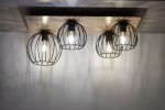 Czarna lampa sufitowa 4-punktowa z oprawami w kształcie siatki, umieszczona na prostokątnej drewnianej podstawie w kolorze dębu. Zbliżenie na źródła światła, które emitują ciepłe światło, tworząc ciekawe cienie na tle.