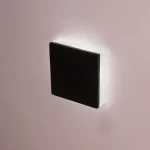 LED oprawa schodowa Lesel 008A L w kształcie kwadratu, w kolorze czarnym z białą ramką, zamontowana na ścianie. Emituje delikatne światło, idealna do oświetlenia schodów lub korytarzy. Ujęcie frontalne.