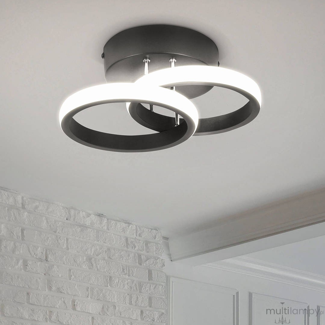 Czarna lampa sufitowa LED 18W z dwoma okręgami, umieszczona na sufitowej podstawie. Okręgi mają matowe wykończenie i emitują białe światło, tworząc nowoczesny efekt. Produkt widoczny na jasnym tle z białymi cegłami.