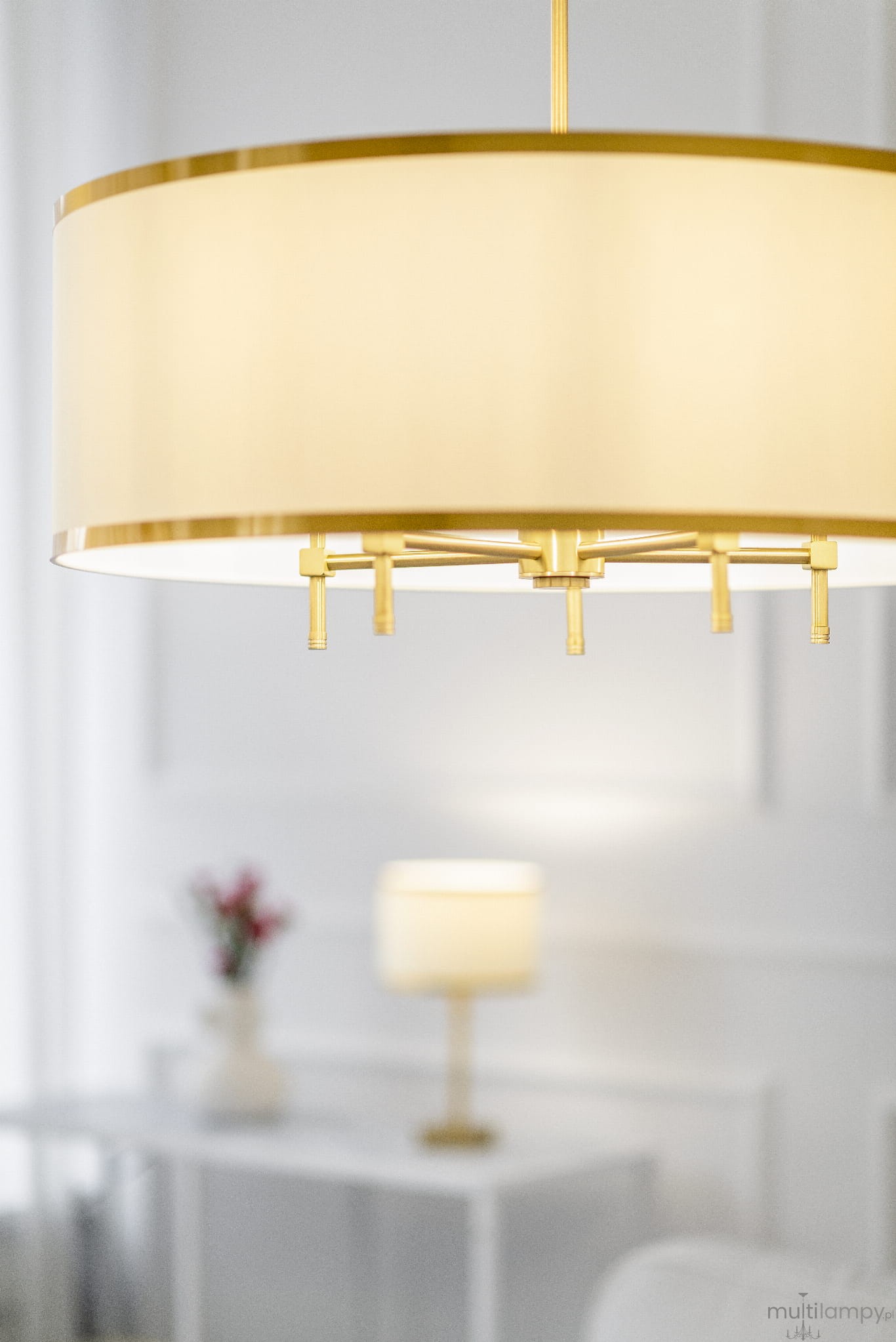 Lampa wisząca Abbano 5-punktowa w kolorze białym i złotym, z okrągłym kloszem i pięcioma metalowymi ramionami. Ujęcie z bliska, ukazujące detale konstrukcji i wykończenia. W tle widoczna lampa stołowa, na białym tle.