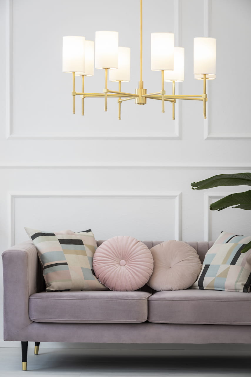 Lampa wisząca Abruzzo 5-punktowa w kolorze białym z metalowymi, złotymi detalami. Posiada pięć cylindrycznych kloszy, zawieszona na długim, złotym ramieniu. Zdjęcie przedstawia lampę nad sofą z poduszkami w pastelowych kolorach.