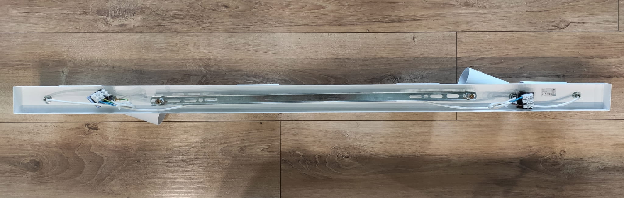 Biała lampa sufitowa (spot) 6-punktowa, widok z góry. Posiada metalową konstrukcję z miejscami na sześć żarówek. Wewnątrz widoczne przewody i mocowania. Prosty, elegancki design, idealna do nowoczesnych wnętrz. Tło drewniane.