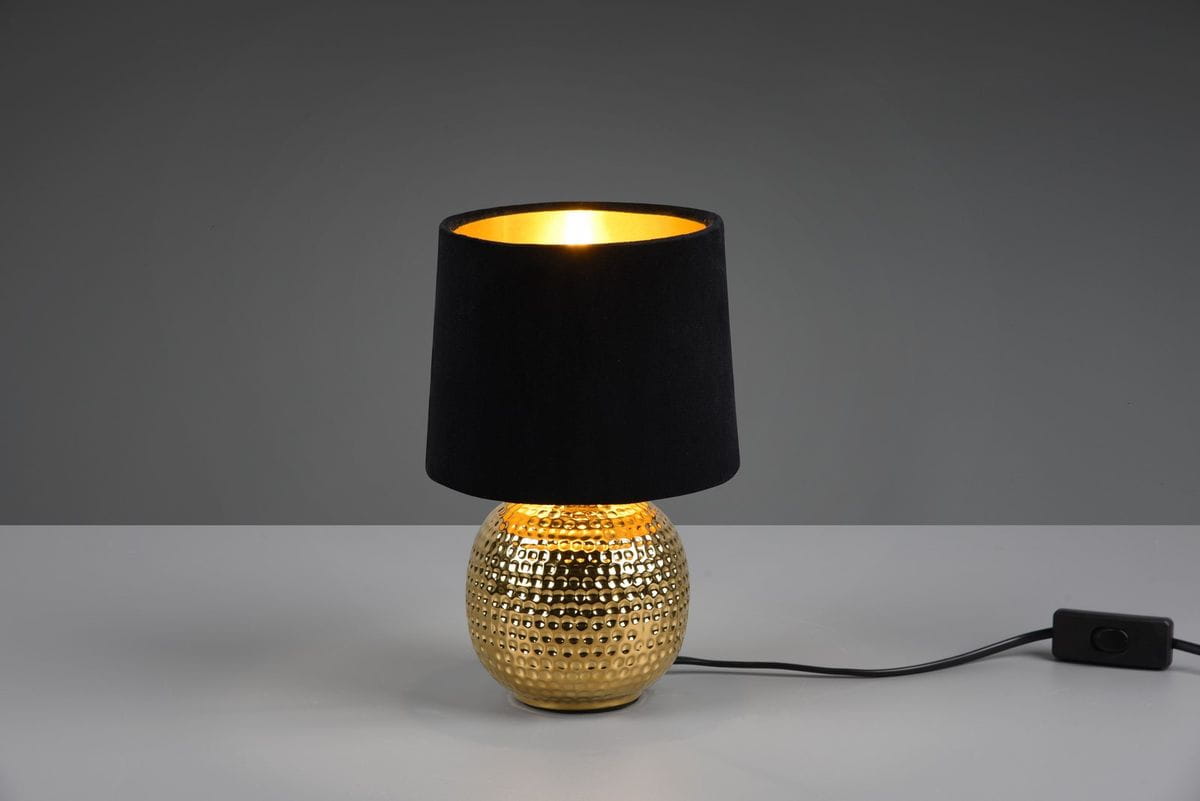 Lampa stołowa Sophia 1-punktowa z okrągłą, złotą podstawą o fakturze w drobne wgłębienia, pokryta czarnym abażurem. Ujęcie z boku, kabel z włącznikiem widoczny na stole. Tło szare.