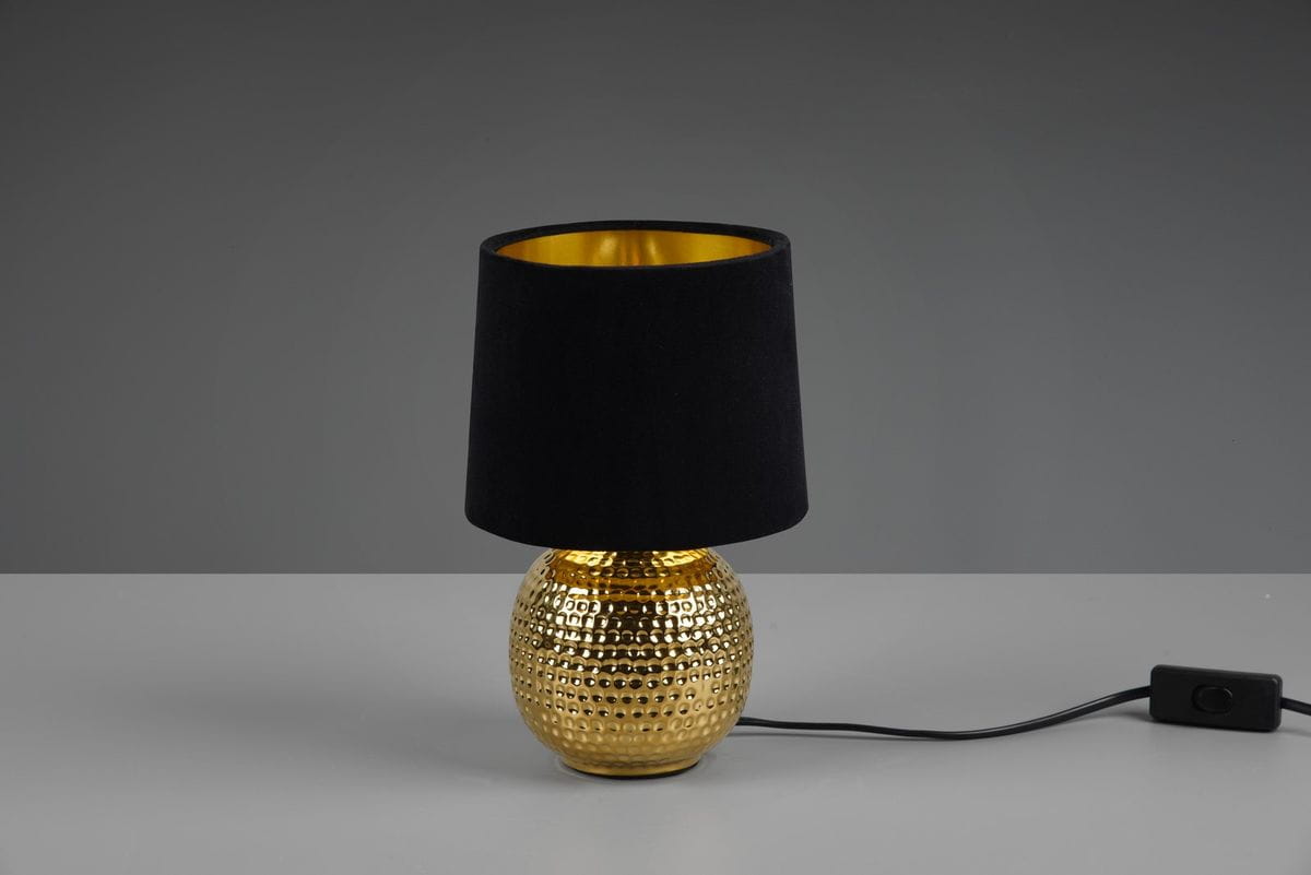 Lampa stołowa Sophia 1-punktowa z okrągłą, złotą podstawą o teksturowanej powierzchni oraz czarnym, materiałowym kloszem. Produkt ustawiony w pozycji stojącej z widocznym przewodem i włącznikiem. Tło szare.