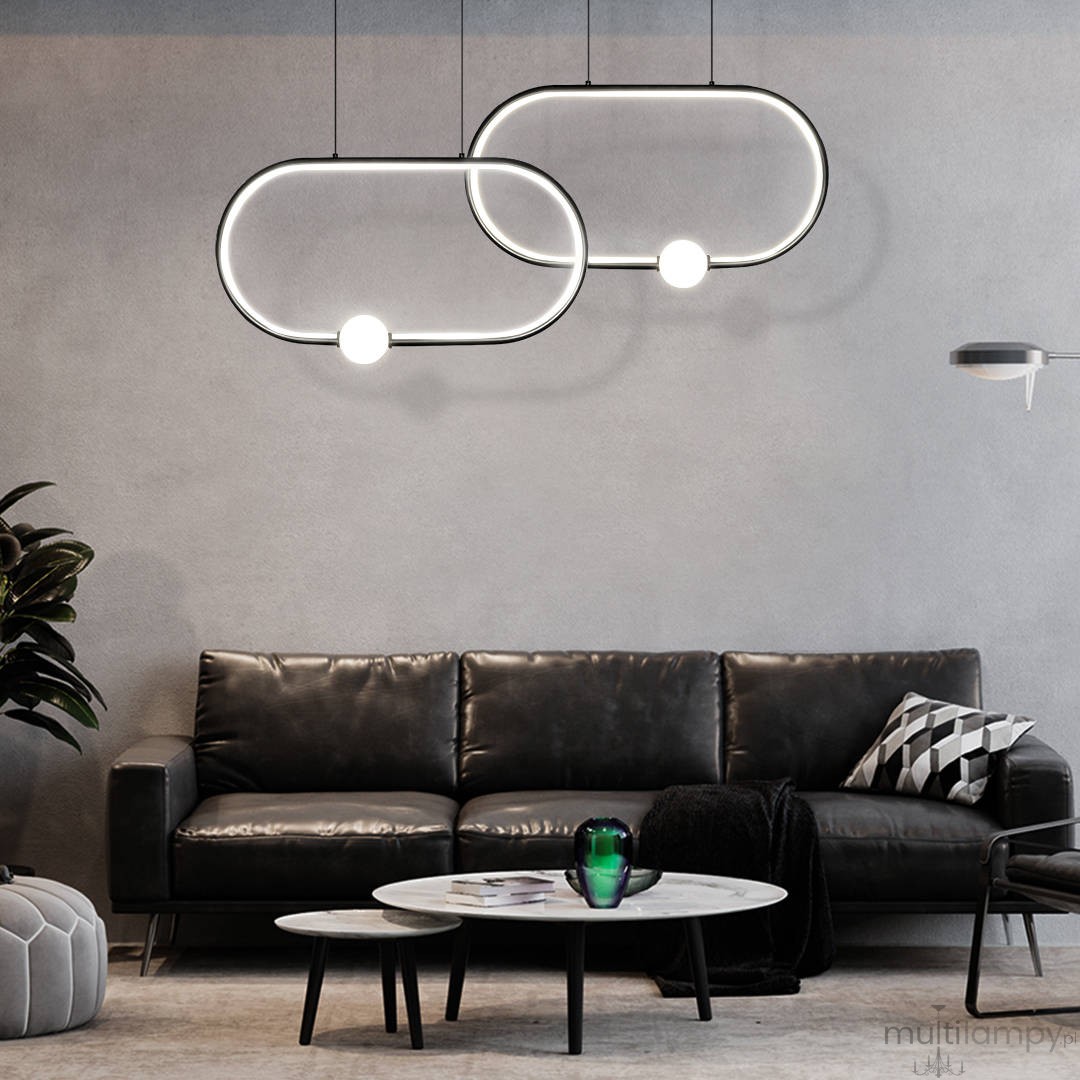 Czarna lampa wisząca Ovo LED 48W, złożona z dwóch okrągłych elementów, zawieszona nad sofą. Lampa emituje światło przez okrągłe źródło, tworząc nowoczesny efekt w aranżacji wnętrza. Tło to szara ściana i ciemne meble.