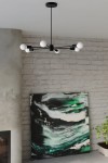 Lango lampa wisząca 6-punktowa w kolorze czarnym, z sześcioma białymi kloszami, umieszczona na cienkich, czarnych ramionach. Lampa zawieszona w przestrzeni, z białą, ceglaną ścianą w tle oraz abstrakcyjnym obrazem w zielono-czarnych odcieniach.