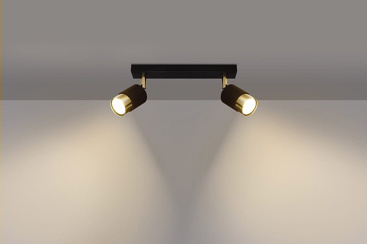 Czarna lampa sufitowa Nero z dwoma punktami świetlnymi. Oświetlenie w formie reflektorów z metalowymi złotymi detalami, zamontowane na prostokątnej podstawie. Ujęcie frontalne, lampa zwrócona w dół, emitująca ciepłe światło na ścianę.