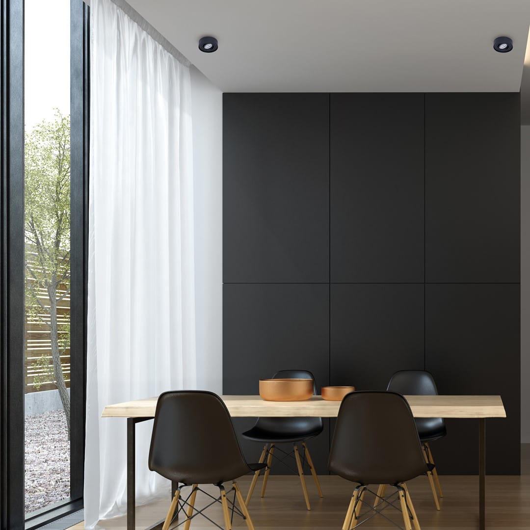 Oprawa do zabudowy Nusa Round 1-punktowa w kolorze czarnym, zamontowana w suficie, o okrągłym kształcie. W tle widoczna jasna zasłona i stół z drewnianym blatem, otoczenie w nowoczesnym stylu.