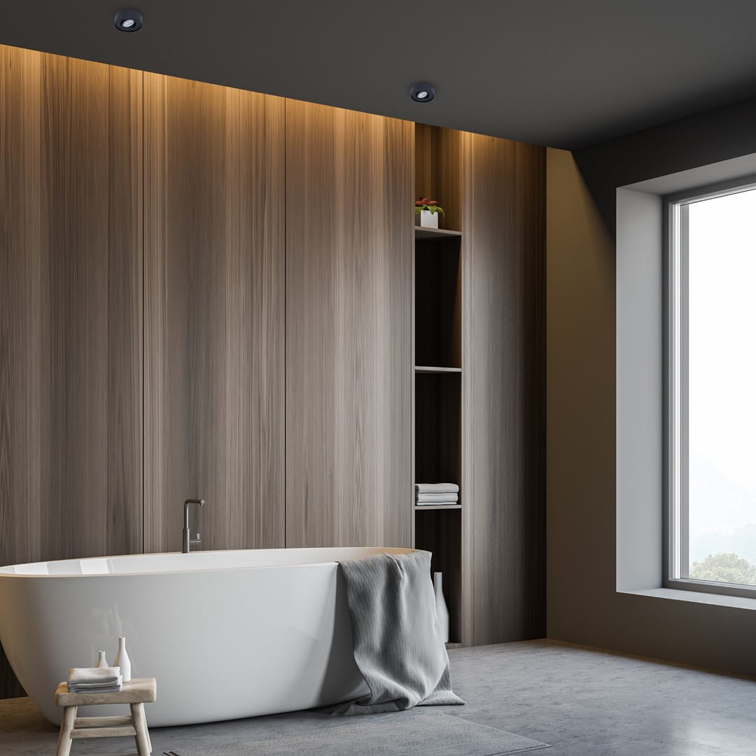 Czarna, okrągła oprawa do zabudowy Nusa Round, umieszczona w suficie w nowoczesnej łazience, z drewnianą ścianą i dużym oknem. Elementy dekoracyjne w tle, takie jak wanna i półka z ręcznikami, tworzą harmonijną aranżację.