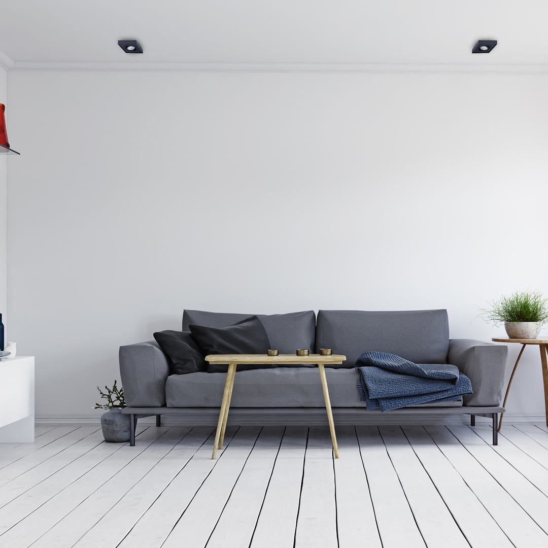 Czarna, kwadratowa oprawa do zabudowy Nusa Square, umieszczona w suficie. W tle nowoczesny salon z szarym sofą, drewnianym stolikiem i jasnymi ścianami. Oprawa o minimalistycznym designie, wkomponowana w jasne, eleganckie wnętrze.