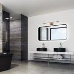 Kinkiet LED Shine IP44 w kolorze różowego złota, montowany nad lustrem. Posiada prostokątną podstawę i emitującą światło oprawę, umieszczony na jasnej ścianie w nowoczesnej łazience z czarnymi umywalkami i dużym lustrem.