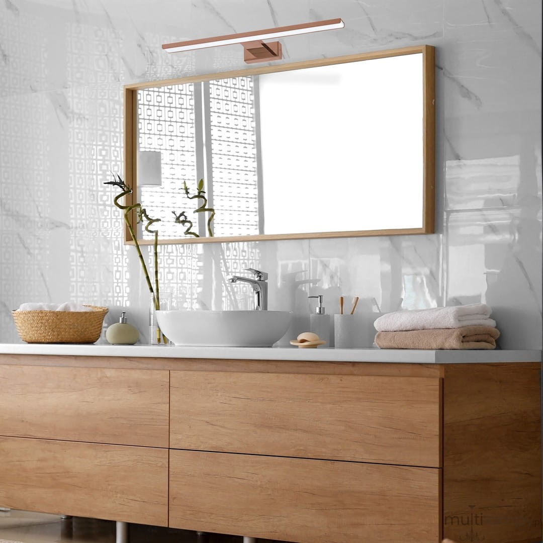 Kinkiet Shine LED IP44 w kolorze różowego złota, zamontowany nad lustrem w nowoczesnej łazience. Oświetlenie ma prostokątny kształt, z metalowym mocowaniem i białą kloszową osłoną. W tle widoczna umywalka, drewniana szafka i ręczniki.