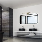Kinkiet Shine LED IP44 w kolorze mosiądzu, 1-punktowy, umieszczony nad prostokątnym lustrem. Produkt o długości 60 cm i mocy 13,8 W, zamontowany na białej ścianie w nowoczesnej łazience z czarnymi umywalkami i szarym tłem.