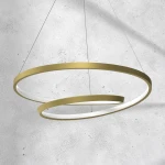 Lampa wisząca Lucero LED 1-punktowa w kolorze złotym, składająca się z dwóch okrągłych elementów, jeden większy i jeden mniejszy, z białym wnętrzem. Zawieszona na cienkich żyłkach, prezentująca nowoczesny design. Tło jest jasne, betonowe.