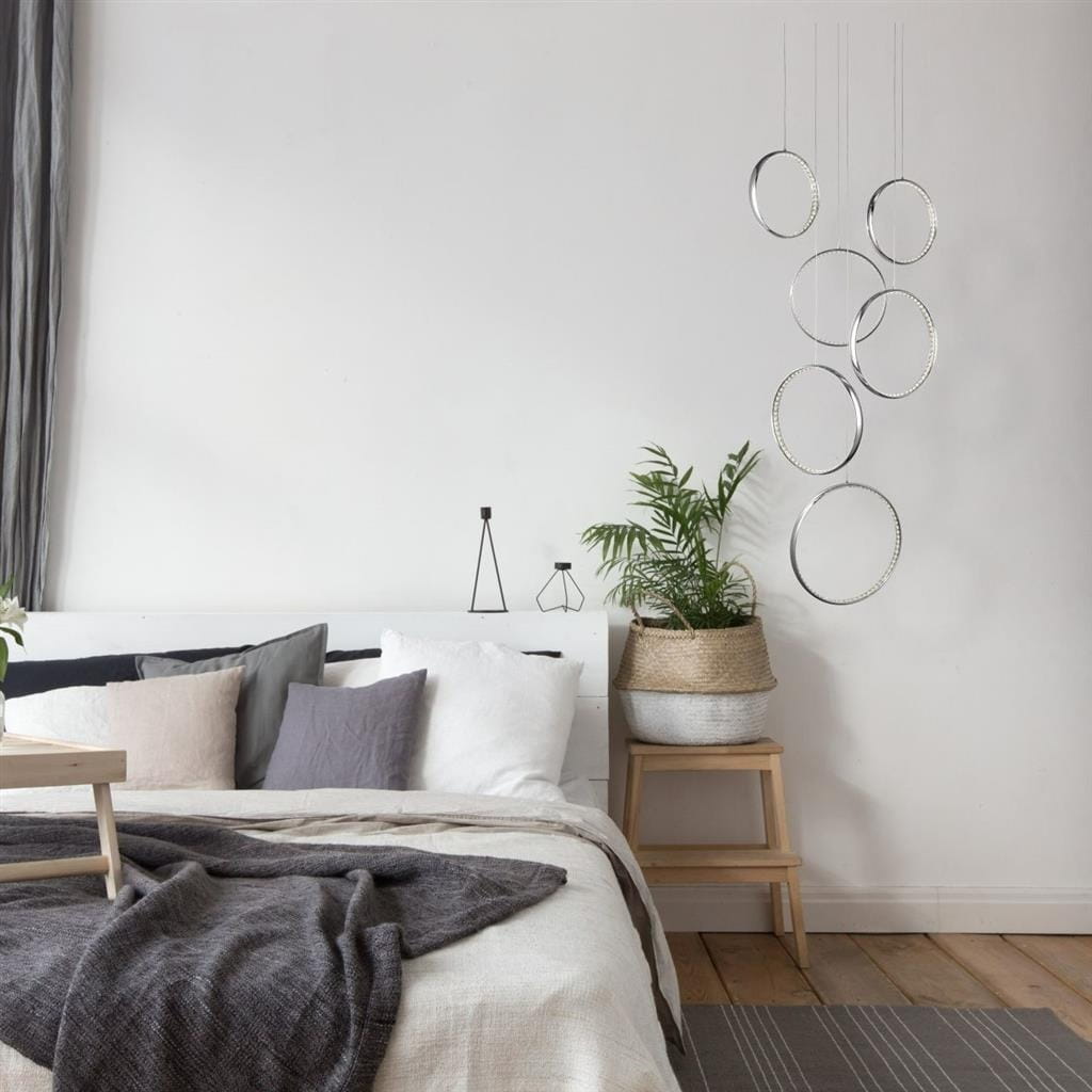 Lampa wisząca Rings LED 6-punktowa w kolorze chromu, składająca się z sześciu okrągłych elementów. Zawieszona nad łóżkiem, w nowoczesnej aranżacji sypialni z rośliną w doniczce i poduszkami w odcieniach szarości.