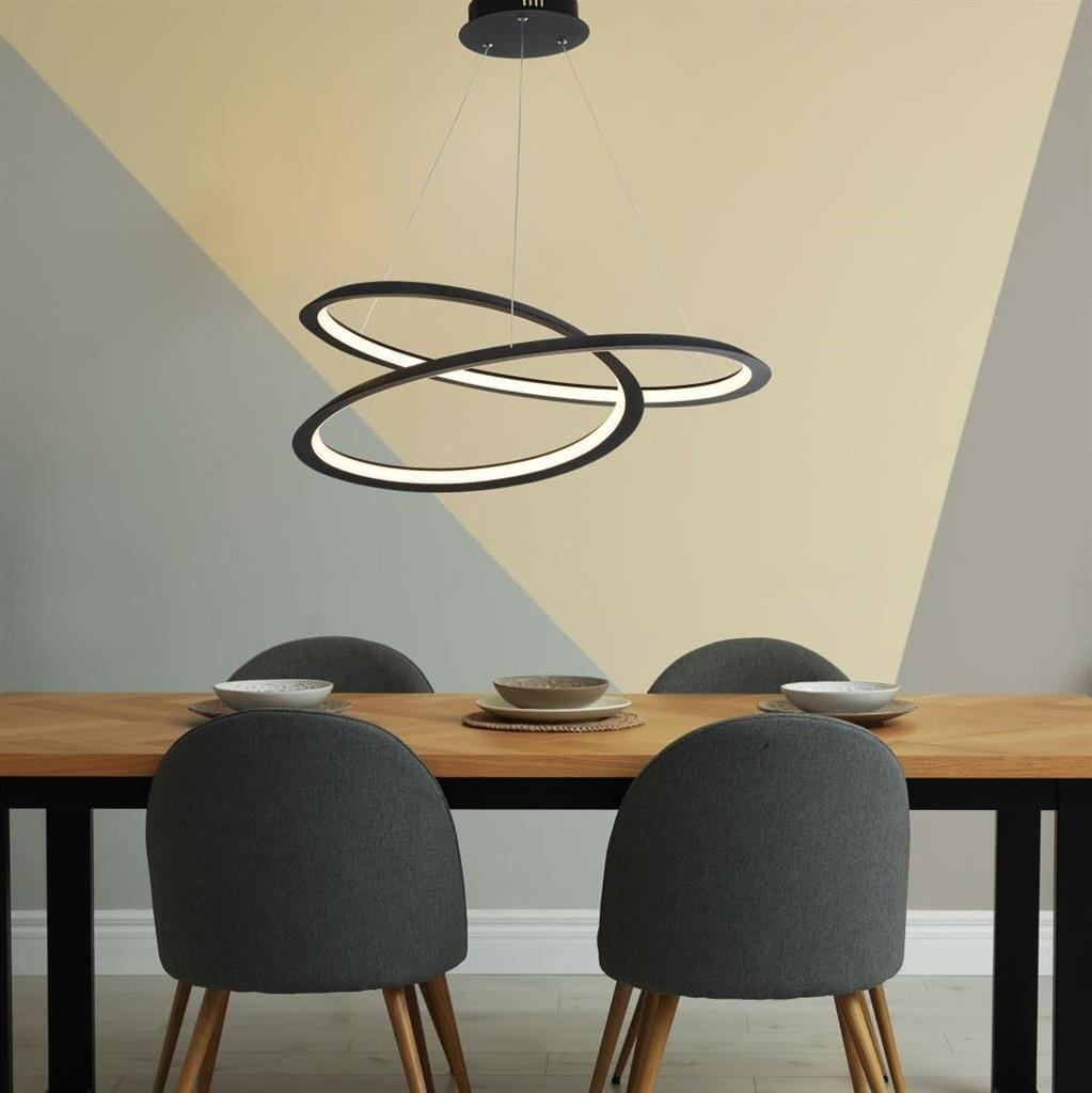 Czarna lampa wisząca Swirl II LED z oprawą w kształcie trzech okręgów, zawieszona nad drewnianym stołem, otoczona szarymi krzesłami. Tło w odcieniach beżu i szarości, podkreślające nowoczesny design.