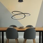 Czarna lampa wisząca Swirl II LED z oprawą w kształcie trzech okręgów, zawieszona nad drewnianym stołem, otoczona szarymi krzesłami. Tło w odcieniach beżu i szarości, podkreślające nowoczesny design.
