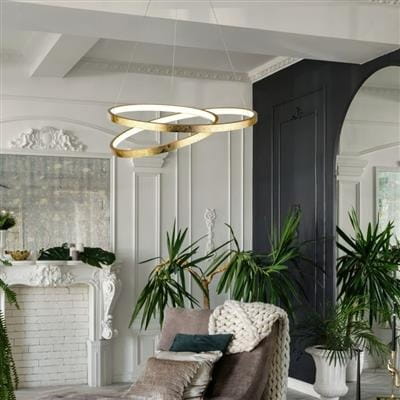 Złota lampa wisząca Float LED, składająca się z dwóch okrągłych pierścieni, wisi w eleganckim wnętrzu z roślinami. Lampa zawieszona na cienkich linkach, w tle widoczny kominek i dekoracyjne elementy architektoniczne.