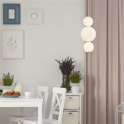 Lampa wisząca Snowball LED w chromowanej oprawie, z trzema okrągłymi kloszami w białym kolorze. Powieszona na tle jasnej ściany, nad białym stołem z krzesłami, w aranżacji z roślinami doniczkowymi i dekoracjami na ścianie.