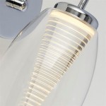Kinkiet Cyclone LED 1-punktowy w chromowanej oprawie. Szklana klosz w formie stożka z dobrze widocznymi, poziomymi żłobieniami, emitujący ciepłe światło. Ujęcie z boku, z widocznym mocowaniem na ścianie.