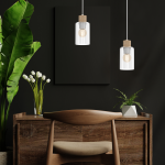 Lampa wisząca Madera 1-punktowa w kolorze białym z przezroczystym kloszem i drewnianym uchwytem. Ujęcie frontalne, zawieszona nad drewnianym biurkiem, w tle czarna ściana z roślinnością.