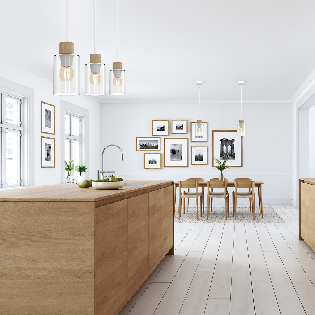 Lampa wisząca Madera 3-punktowa w kolorze białym z przezroczystymi kloszami i drewnianymi detalami, zawieszona nad drewnianym blatem kuchennym. W tle widoczne zdjęcia w ramach i stół z krzesłami.