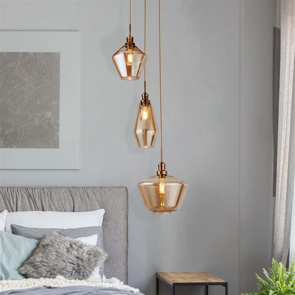 Lampa wisząca Mia 3-punktowa w kolorze antycznego mosiądzu z trzema szklanymi kloszami o różnorodnych kształtach, zawieszona nad łóżkiem w jasnym wnętrzu. Klosze mają ciepłą, przezroczystą barwę, a całość wisząca na długich kablach.