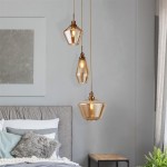 Lampa wisząca Mia 3-punktowa w kolorze antycznego mosiądzu z trzema szklanymi kloszami o różnorodnych kształtach, zawieszona nad łóżkiem w jasnym wnętrzu. Klosze mają ciepłą, przezroczystą barwę, a całość wisząca na długich kablach.
