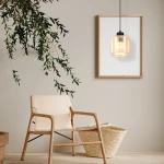 Lampa wisząca Sombra 1-punktowa w kolorze czarnym z bursztynowym kloszem, zawieszona na czarnym kablu. Otoczona nowoczesnym wnętrzem z drewnianym krzesłem, koszem i roślinnością w tle.
